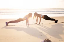 Exercícios para fazer na praia no verão: Dicas imperdíveis Two women doing push-ups on a sandy beach during sunset for a fitness workout.