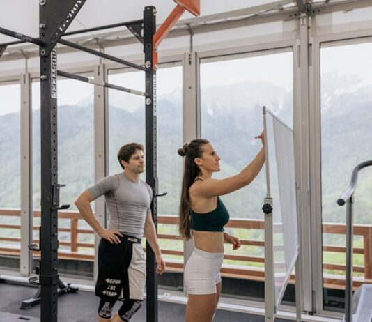 Plano de treino para o Verão: Surpreenda-se com os resultados A gym coach instructs on whiteboard beside fitness equipment with mountain view.