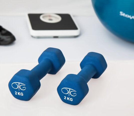 Treino em casa nos dias de chuva: Transforme o seu espaço Blue dumbbells placed on a white surface with fitness equipment in the background.