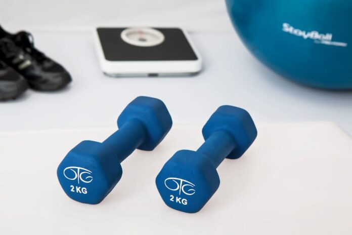 Treino em casa nos dias de chuva: transforme seu espaço! Blue dumbbells placed on a white surface with fitness equipment in the background.